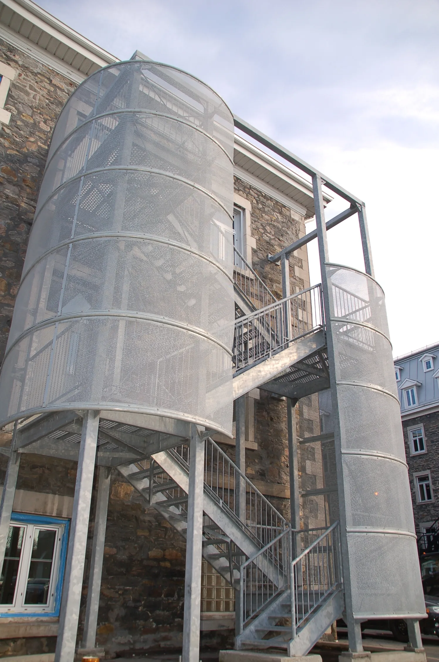 Escalier extérieur en acier galvanisé, avec paliers et cages en métal perforé, adossé à un bâtiment de pierre